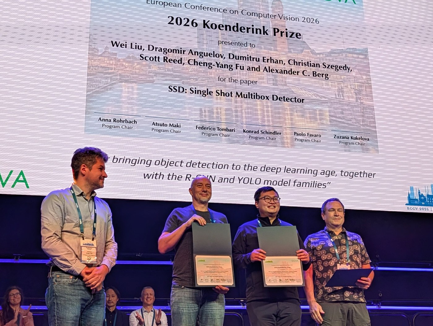 Four men on stage at ECCV 2026 in front of a large slide announcing the 2026 Koenderink Prize for SSD: Single Shot MultiBox Detector. Two of them hold open award certificates.